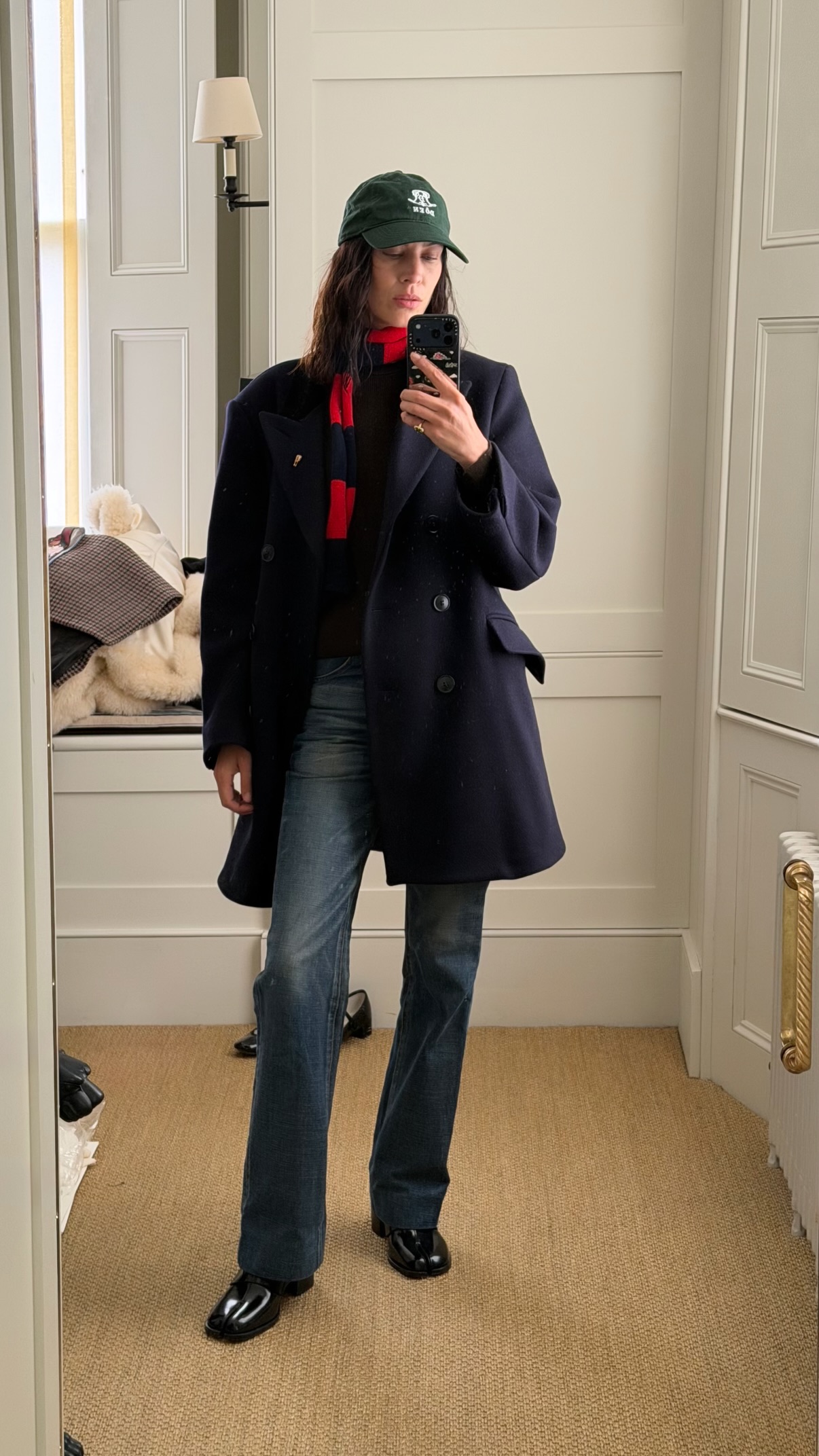 Alexa Chung takes a mirror selfie wearing a navy coat with stove pipe jeans, Tabi loafers and a green cap.