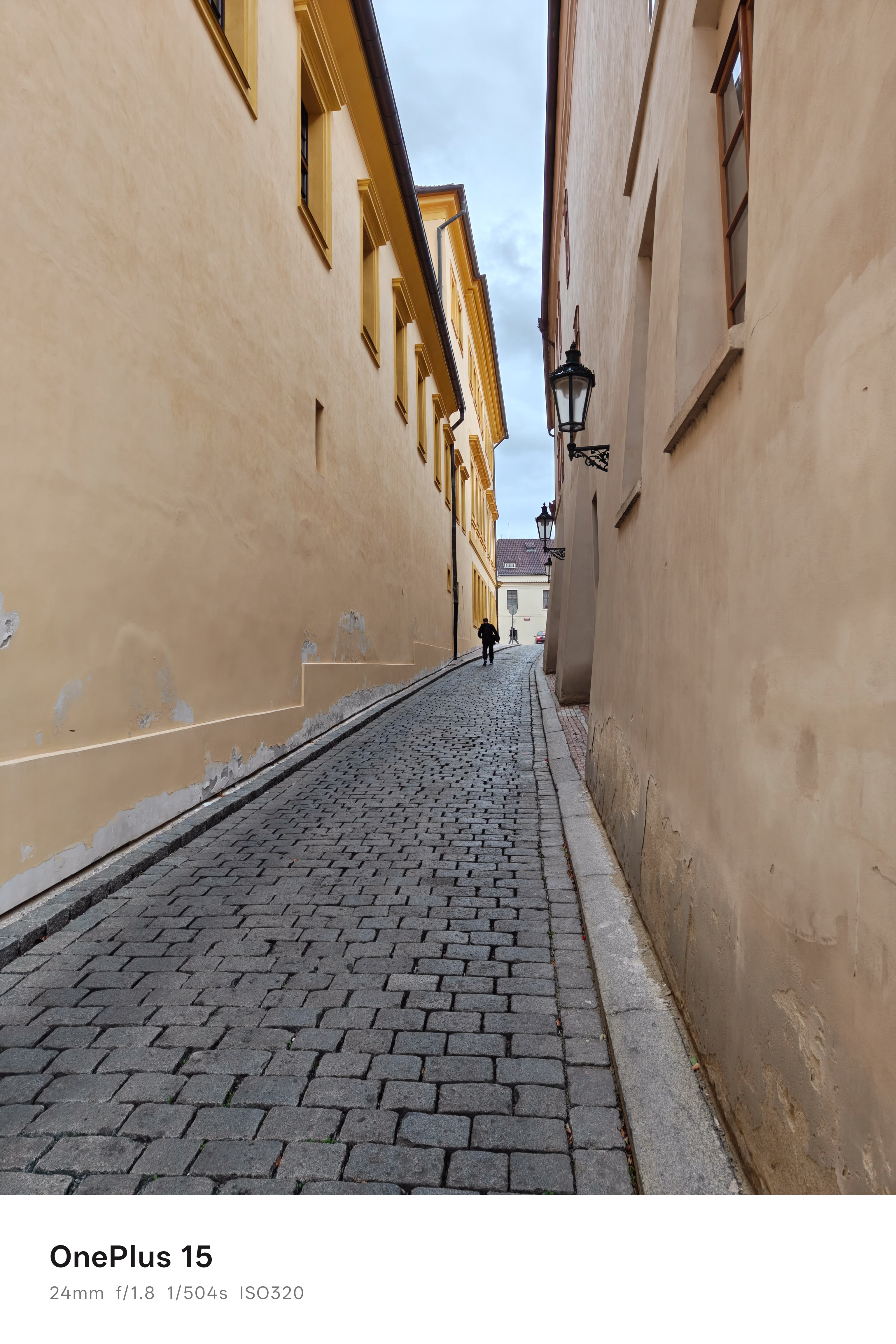A person walking up a long narrow cobbled path between two building buildings in Prague