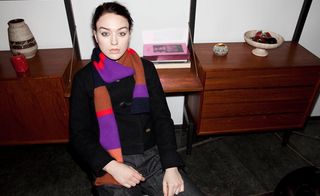 Female model sat in a chair, wearing black jacket, dark trouser, multicolour stripe scarf, dark wood unit with ornament =s and vases on the top, white wall with black frame, dark grey floor