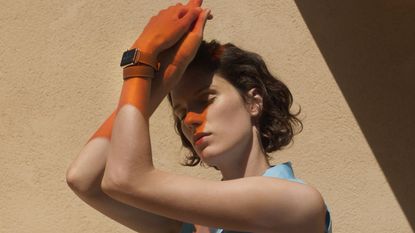 A woman stands with her arms entwined and raised above her head to shade her face and displaying an apple watch on her wrist.