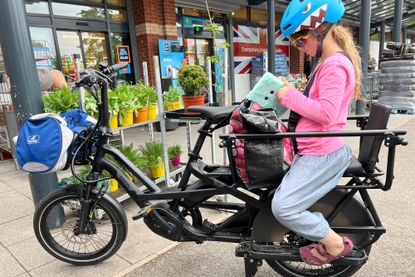 The best electric cargo bikes can replace the car when going to the shops like this image which shows a child wearing a pick top in a blue helmet on the back of a Tern GSD S10 LX which has bags front and back in front of a busy supermarket