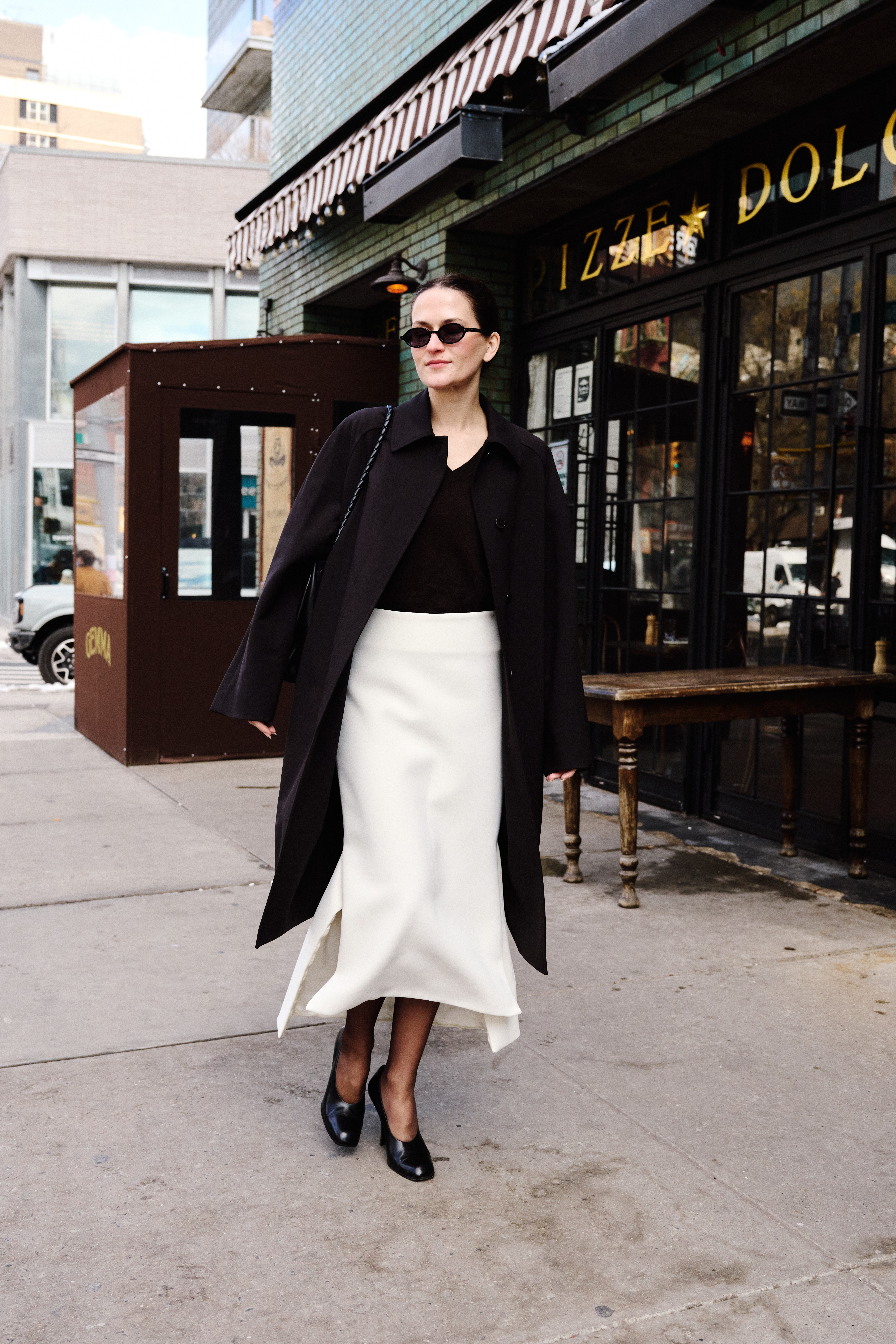 Eliza Huber wearing a brown car coat, brown cashmere sweater, cream split-hem skirt, brown tights, and black heels while carrying a black Bottega Veneta Ciao Ciao bag outside of the Bowery Hotel.