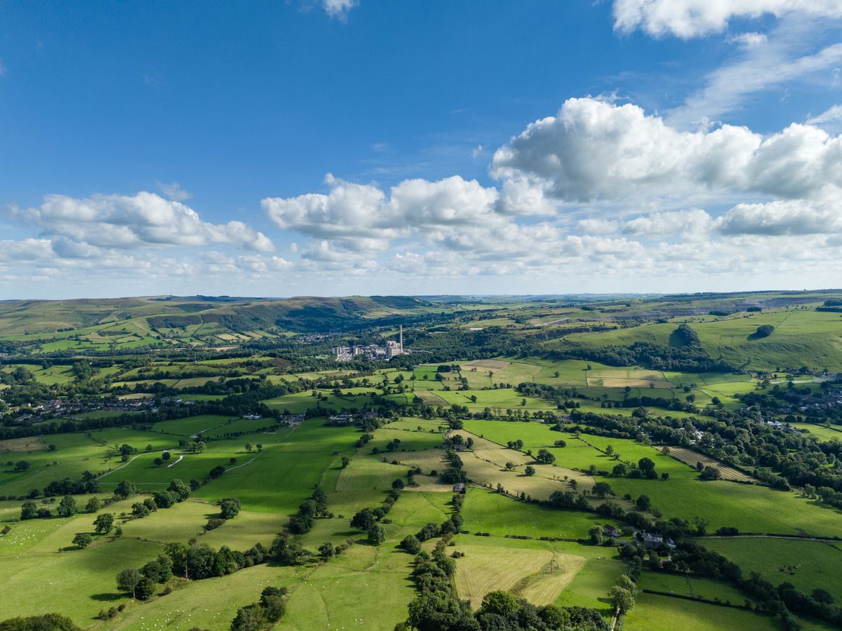 Image taken by DJI Air 3 (a camera drone). In the distance you can see an industrial plant. It is surrounded by dark green trees. In the foreground are rolling green hills dotted with dark green trees and above is a blue sky dotted with white, fluffy clouds.