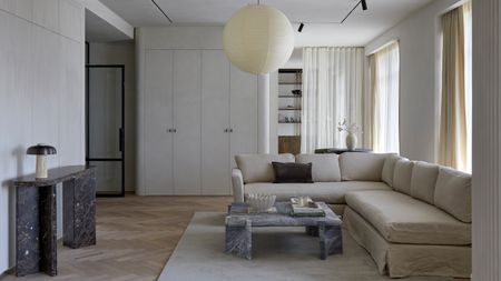 Living room with white walls and built-in cupboards, round paper pendant light, blue-gray marble coffee table, gray-brown marble console table, cream corner sofa and cream rug