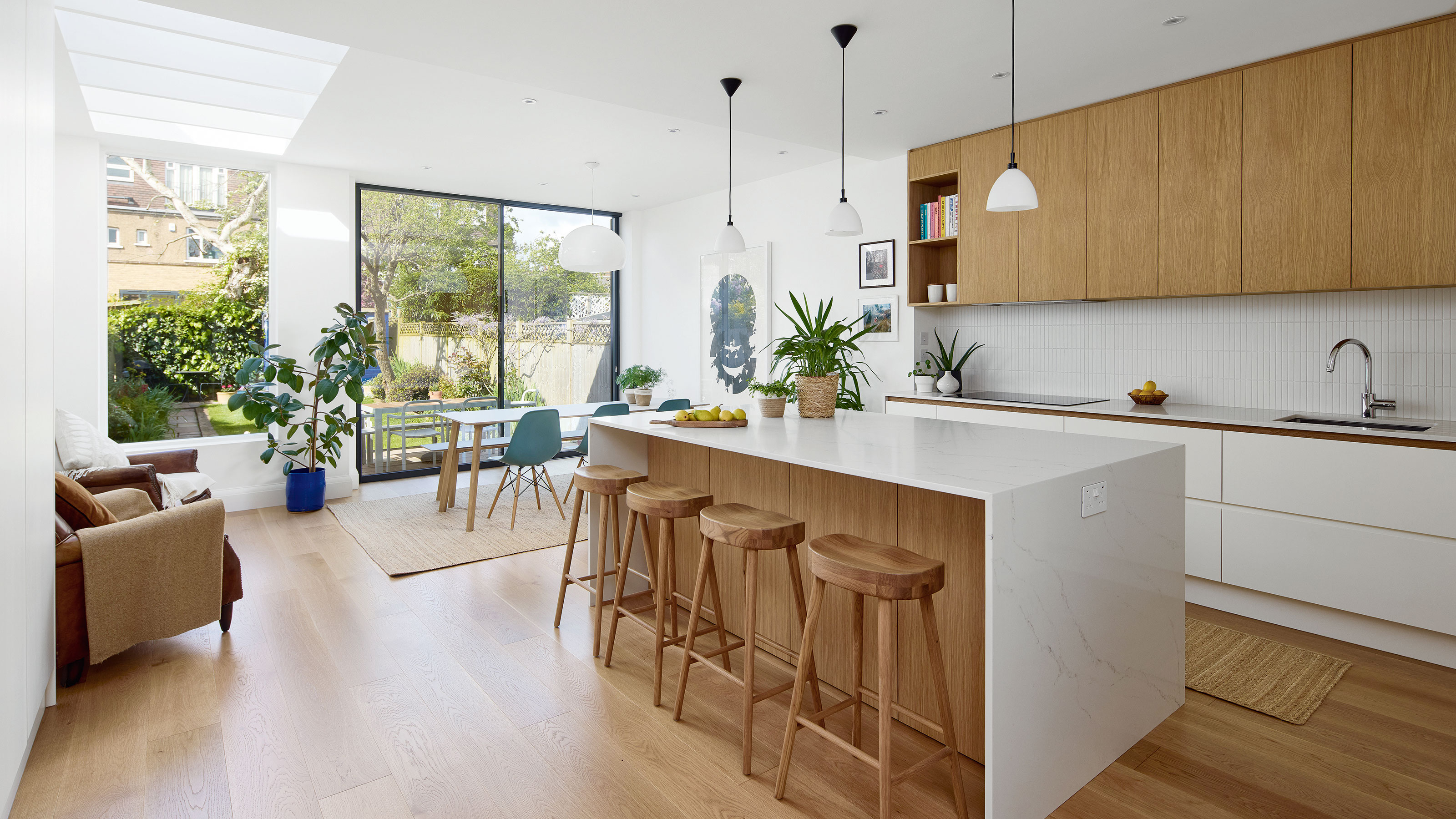 White kitchen with wooden floor, sleek cabinetry and white island with wooden bar stools. Skylights in roof and floor-to-ceiling window out to garden