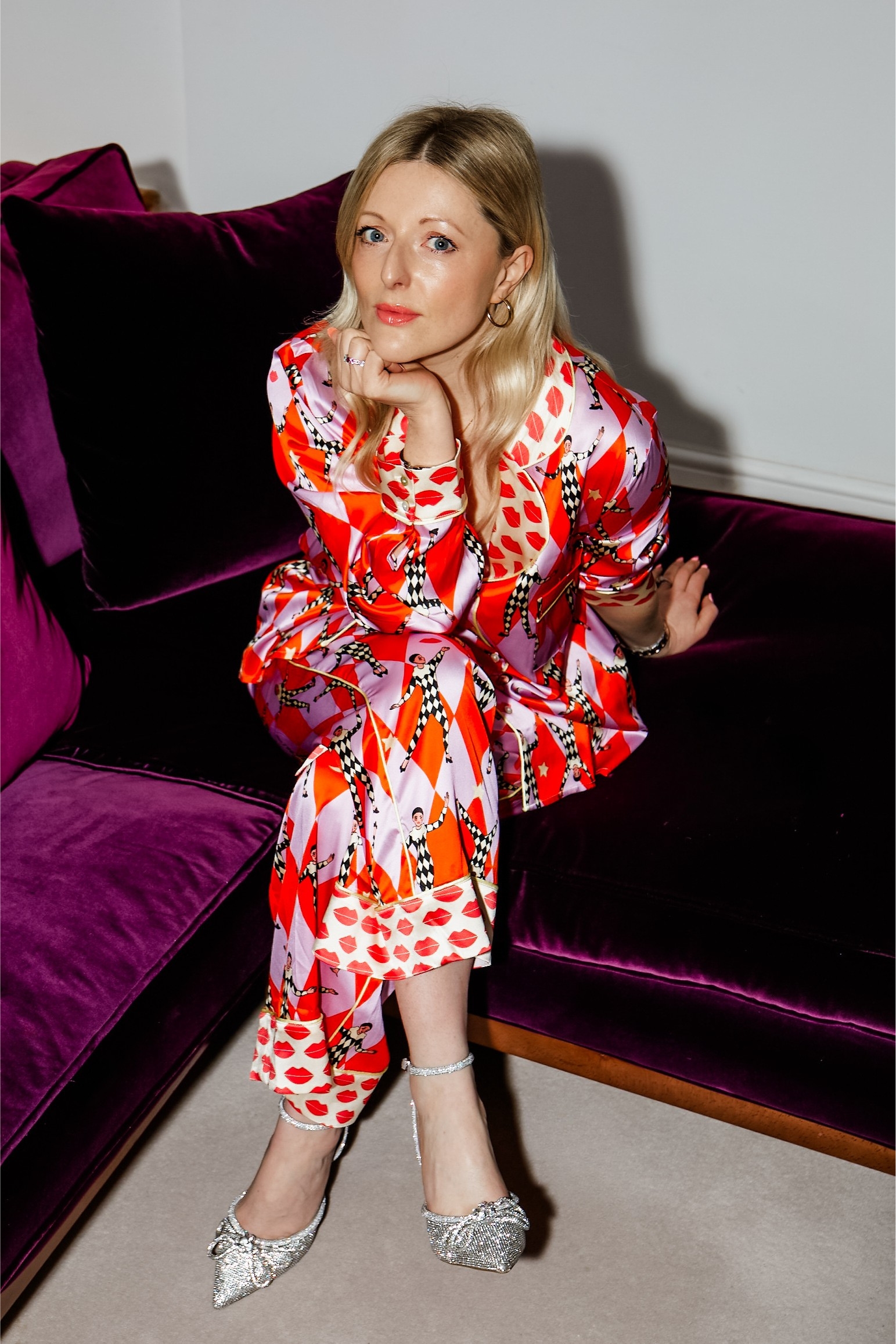A woman sits on a burgundy sofa wearing a red and pink patterned silk suit