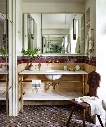A Mediterranean bathroom with a gold and marble vanity, tiled mosaic floor, zellige tiles in beige and burgundy on the walls, and a large vanity mirror