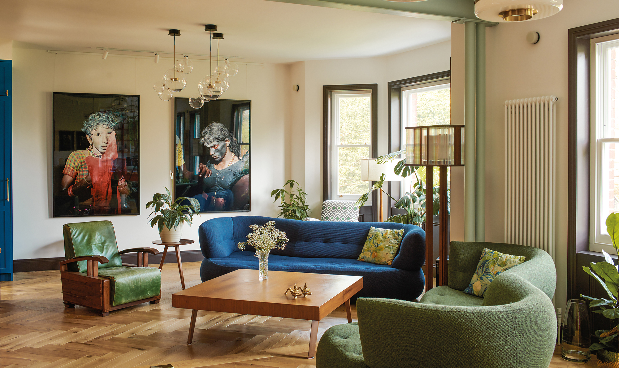 modern living room with blue sofa and green sofa and armchair with timber floors, large artworks, square timber coffee table, radiator on the wall, floor lamp, and pendant lights