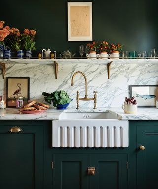 dark green kitchen with marble countertops and backsplash, fluted white sink, brass hardware, marble shelf with flowers, glassware, artwork