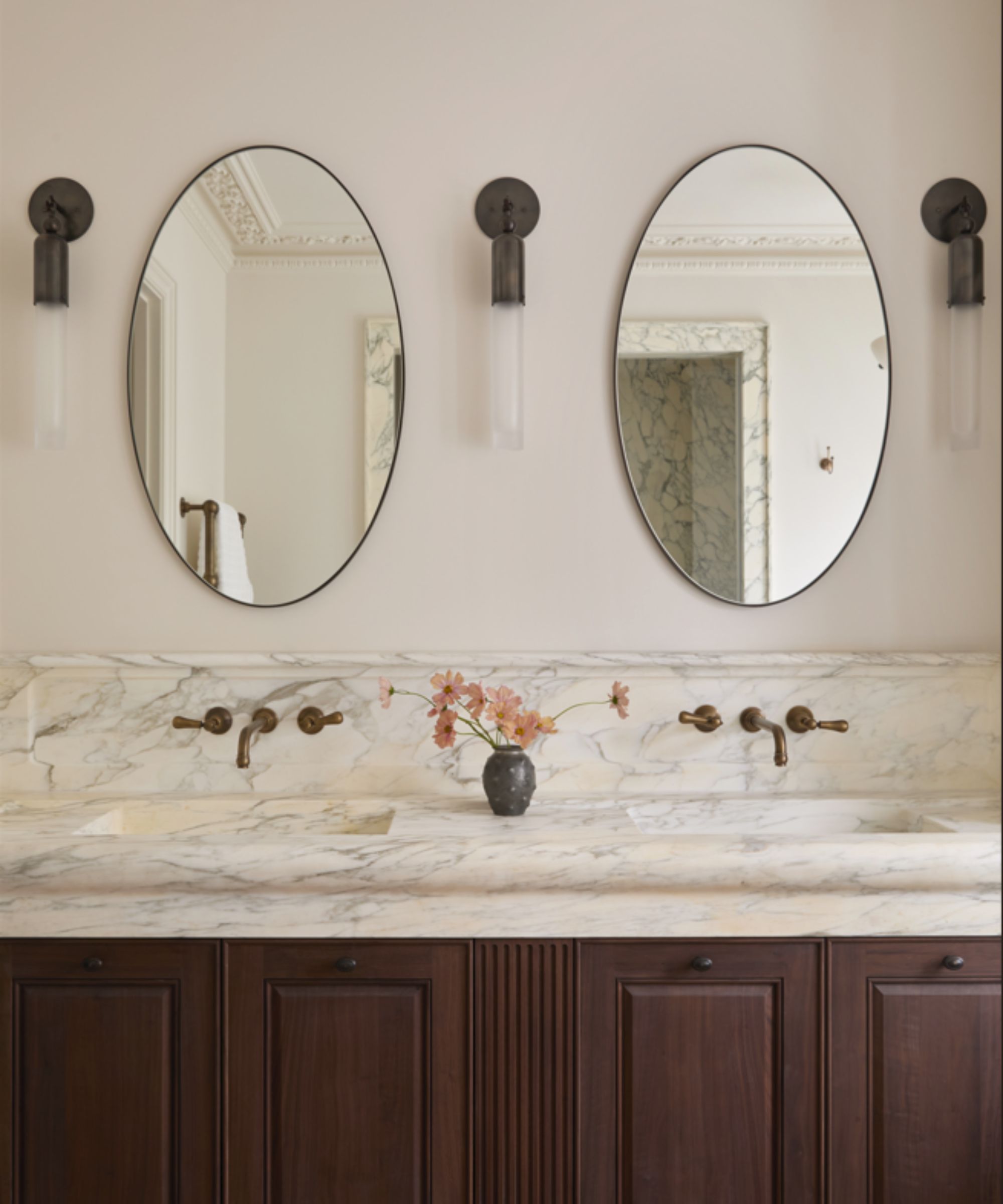 Double vanity area in bathroom with wooden cabinetry, two marble sinks with backsplash, vase of flowers in middle, two oval mirrows hanging above with three wall lights in-between