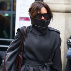 Victoria Beckham wears a high neck trench coat with oversize sunglasses and a tote bag