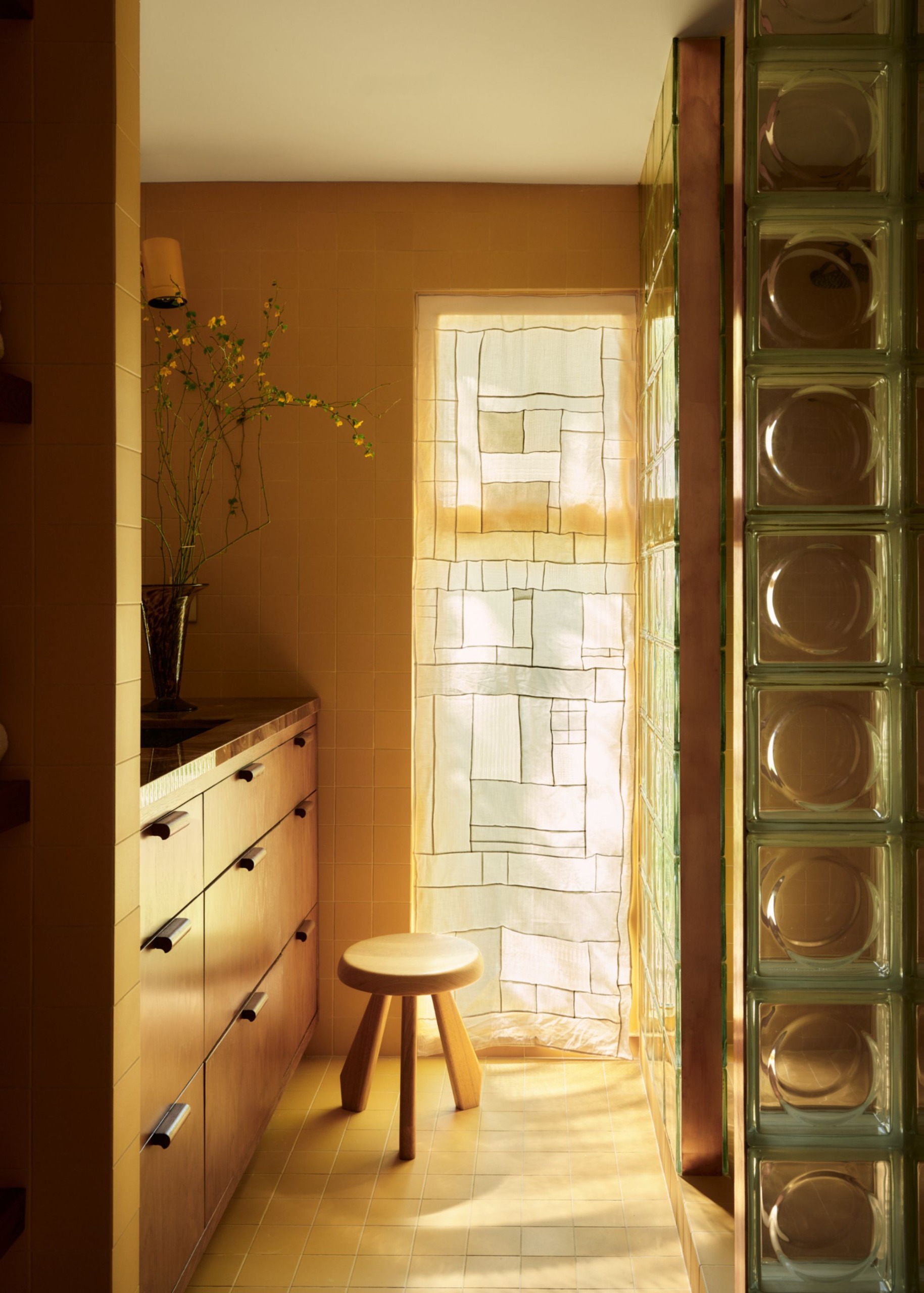 A modern bathroom with ochre wall and floor tiles, a narrow floor-to-ceiling window with a geometric patterned sheer window treatment, a glass brick shower wall, and a wooden vanity.