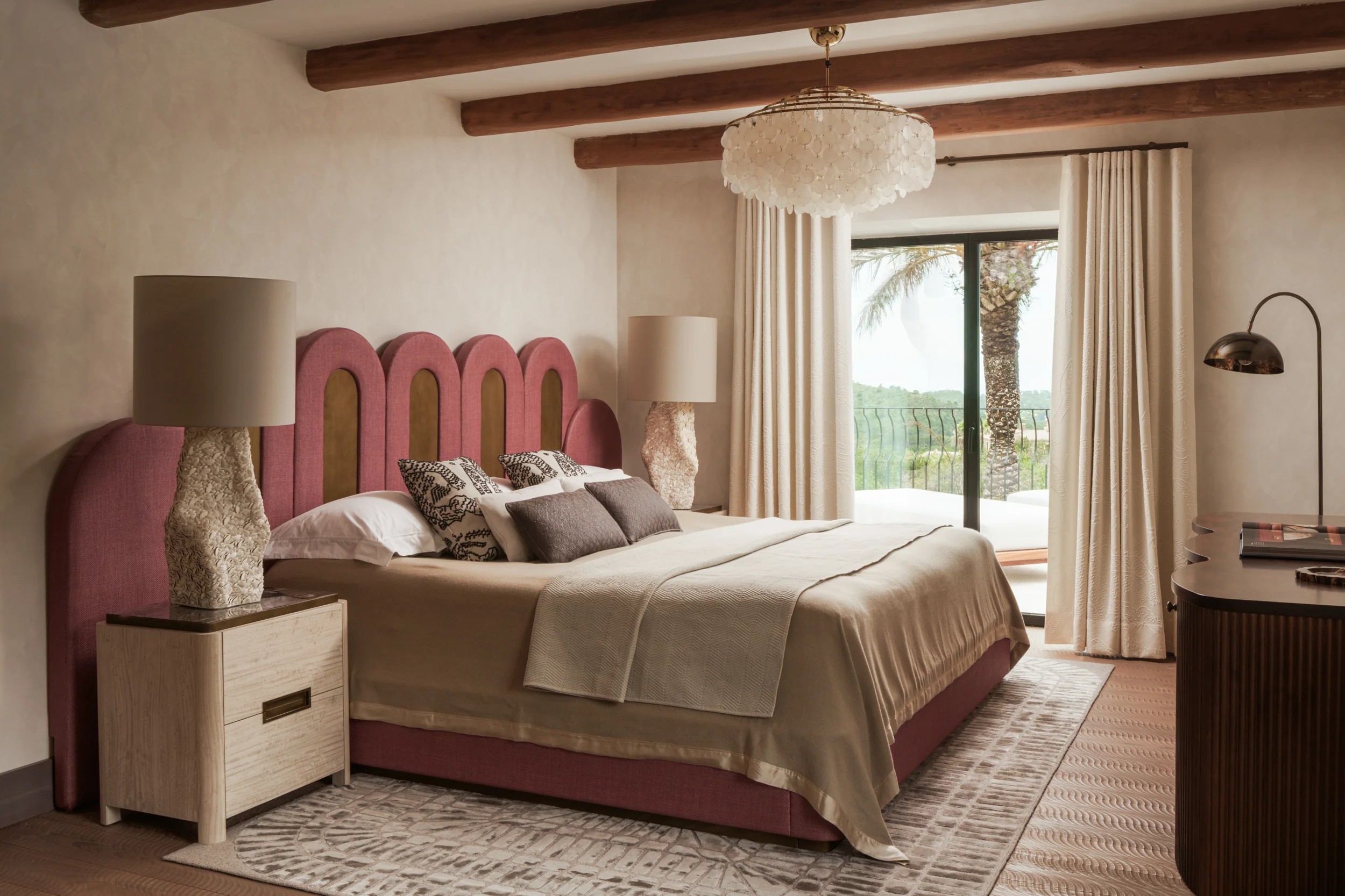 A coastal bedroom with wooden floors, layered beige textiles, sculptural lighting, and garden views.