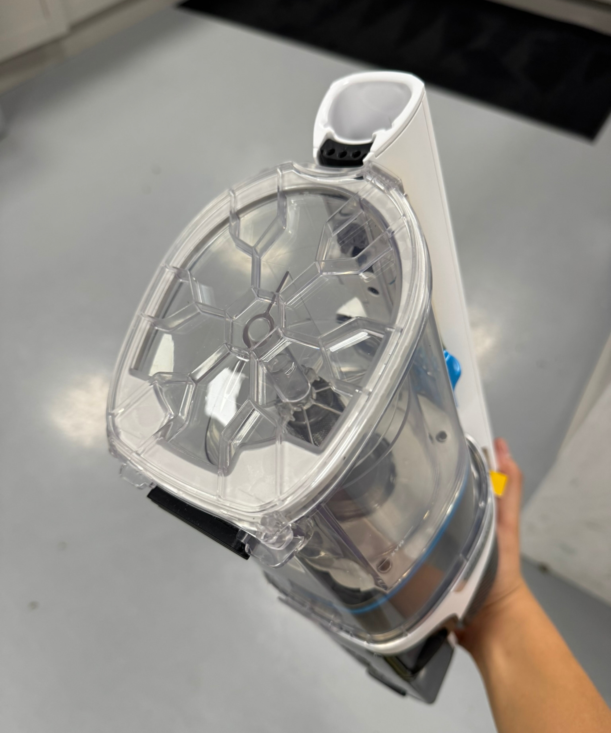 Hand holding the Shark PowerPro Pet cordless vacuum, revealing see-through dustbin, with gray floor in background.