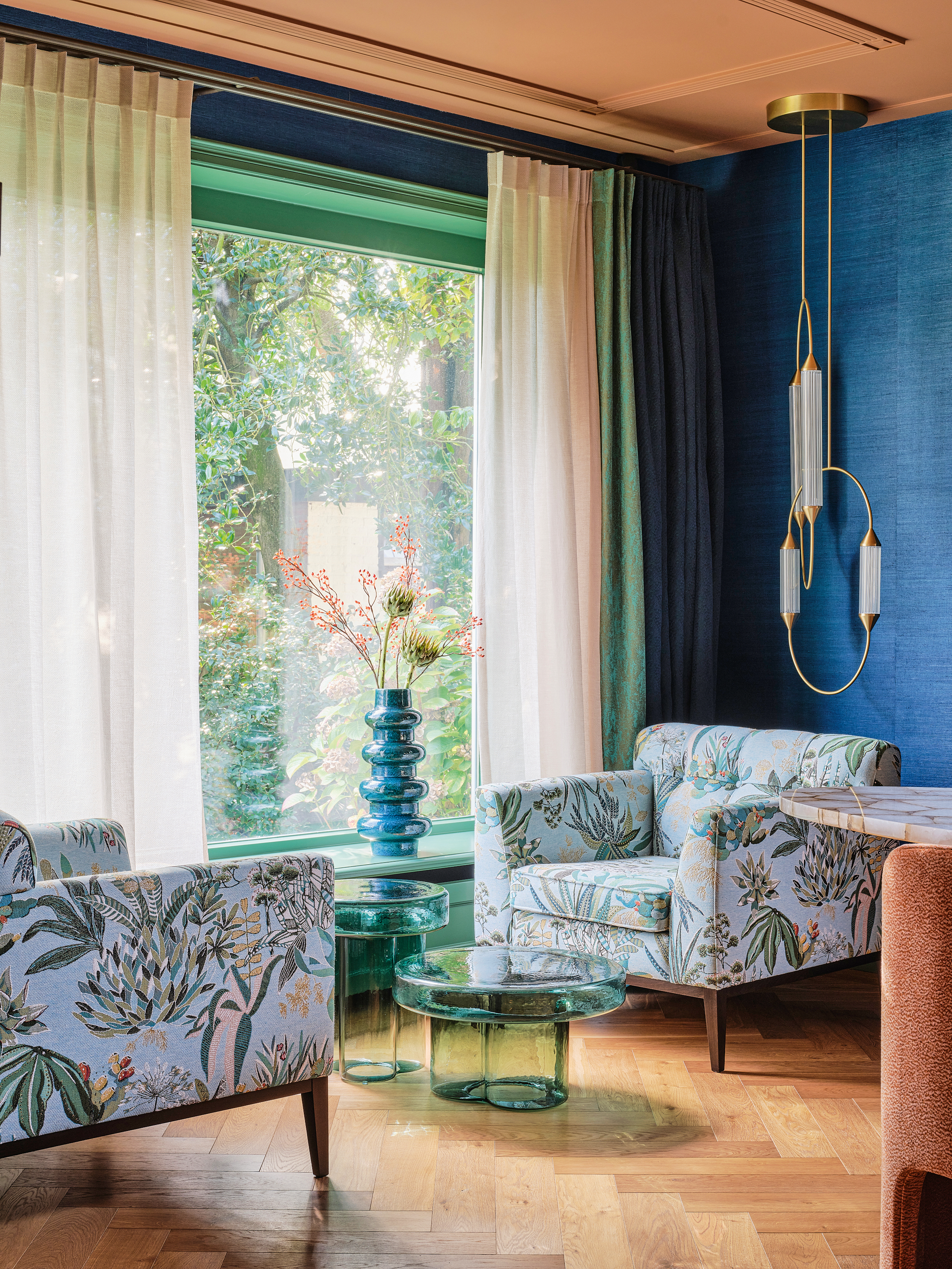Corner of a dining room with bright blue walls, green and cream sheer curtains, pale blue floral armchairs, green glass coffee tables and a blue vase