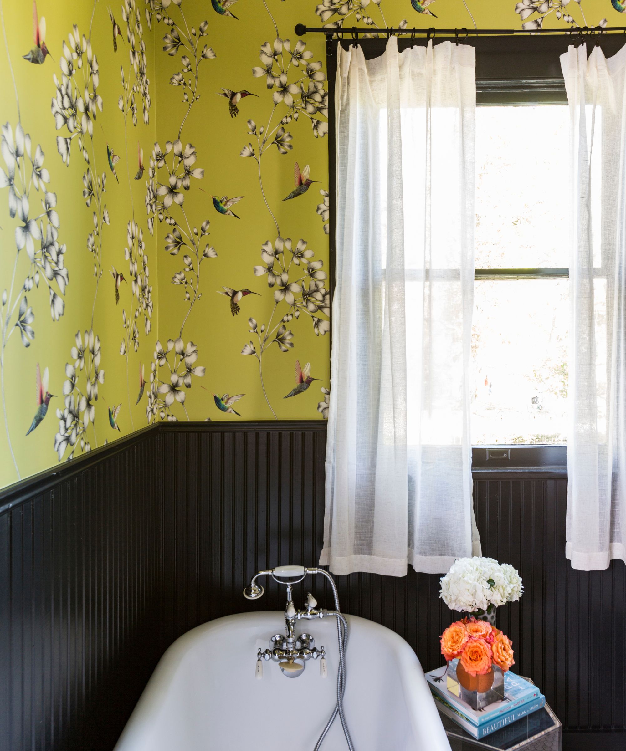 A bathroom with yellow floral wallpaper and black paneling on the lower walls. Sheer white window curtains and a white roll-top bath.