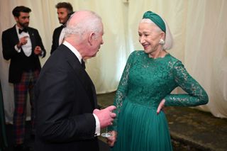 Helen Mirren wearing a green lace dress standing with her hand on her hip talking to The King, wearing a tux