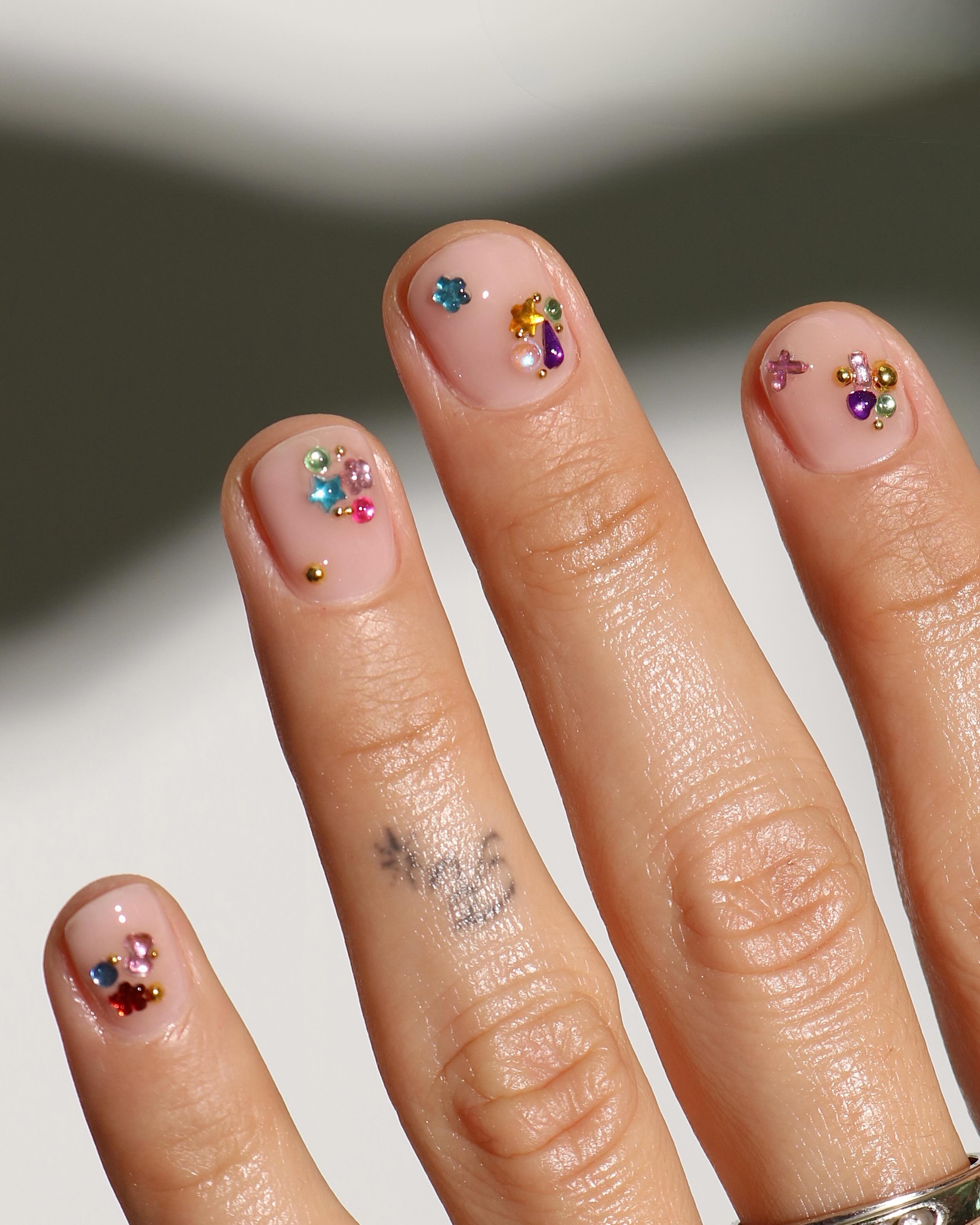 Short nude nails decorated with scattered colorful gems, miniature floral charms, and small blue stars