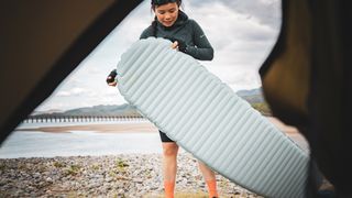 A woman stands outside a tent, viewed from inside, holding a Therm-a-Rest NeoAir XTherm NXT Sleeping Pad on a stony beach.