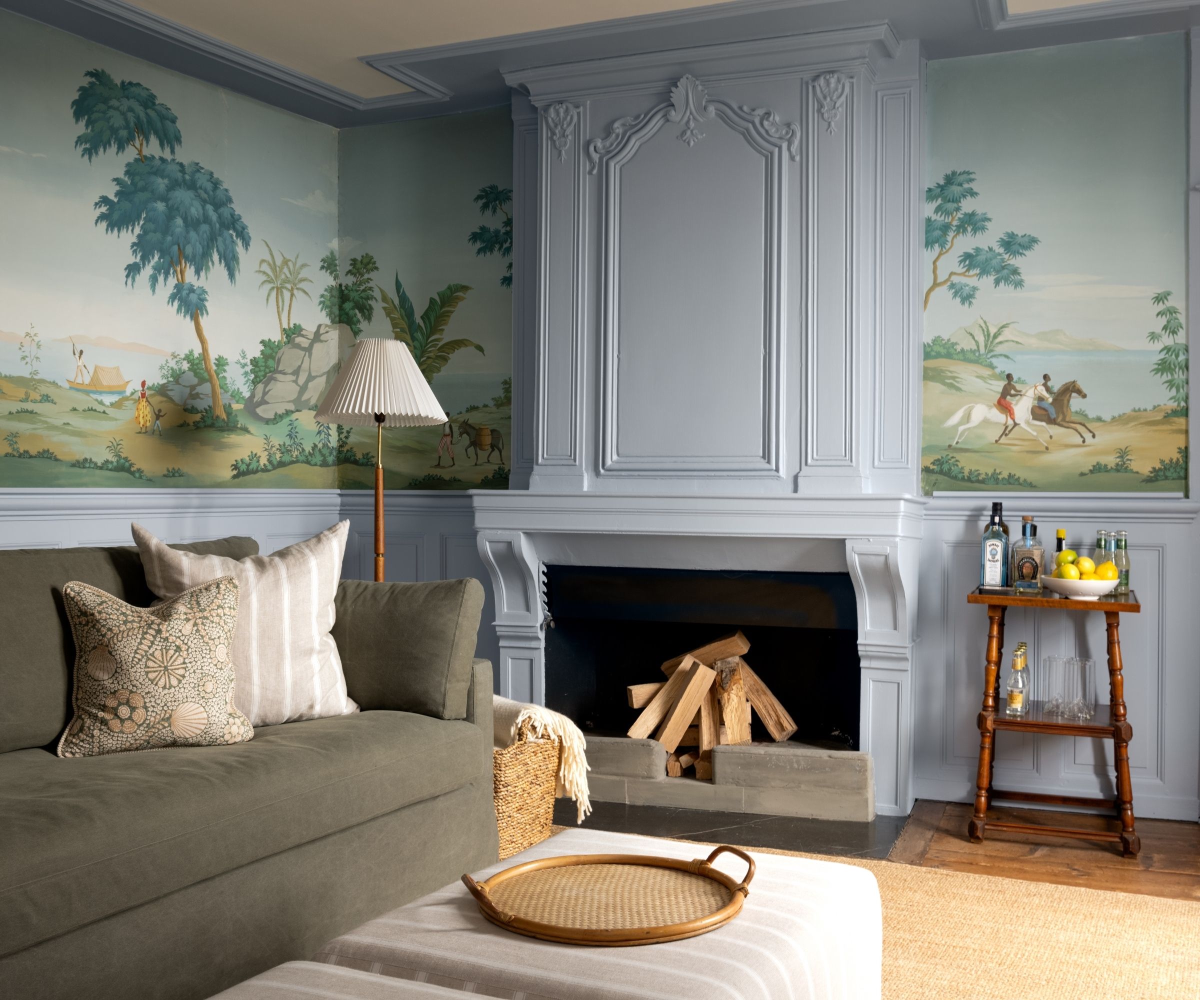 Living room with pale blue painted ceiling trim and fireplace, a mural landscape wallpaper, a dark green sofa, a large fireplace with logs, two striped ottomans, and a drinks table in the corner.
