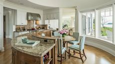 Bright coastal home with rounded kitchen features and layout. There is a marble island in a semi circle, striped blue and white upholstered chairs around a round table and bright tall windows with the ocean in view