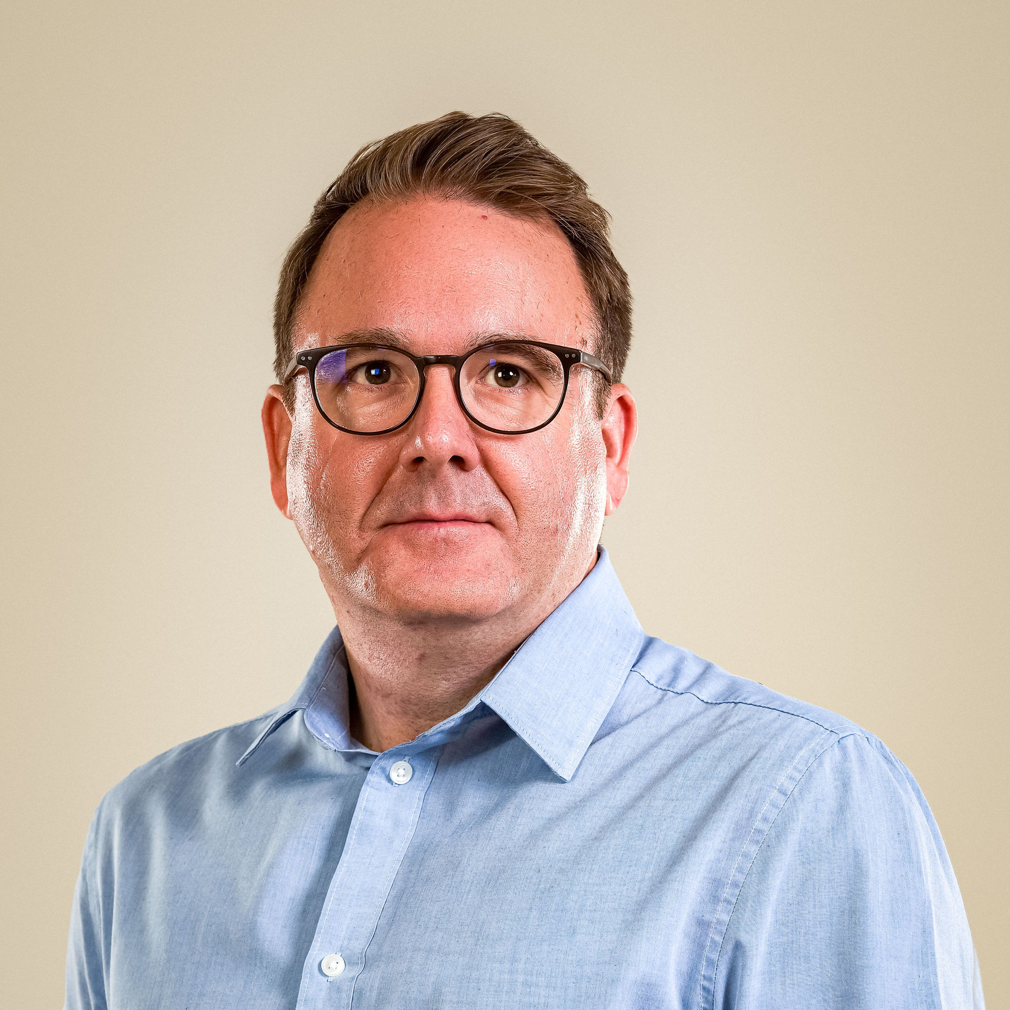 male with short brown hair and black framed glasses wearing blue shirt stood against cream background