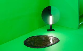 A dark grey rug with the appearance of a puddle of water sits on a bright green floor. A large scale circular lamp stands on the floor behind against a bright green wall.