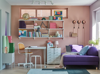 a multi coloured home office in living room space, with a pink wall, a white shelving unit, a small purple armchair, and a white desk and drawers