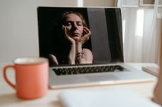 Portrait of a woman sitting at the notebook, red cup of tea on the table. Reflection of a face in the monitor screen, chin on hands. She looks tired.