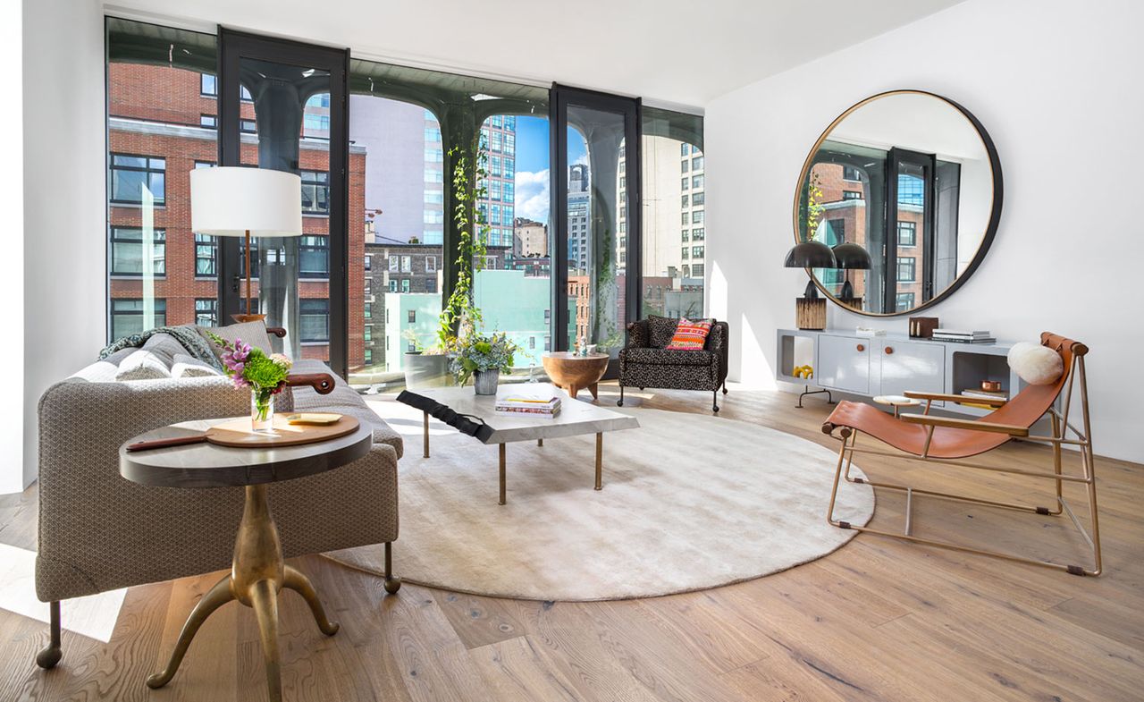 A lounge area with a light brown sofa, a leather chair, a black fabric chair, a coffee table, a white round rug, a grey console, a round wall mirror and sliding doors.