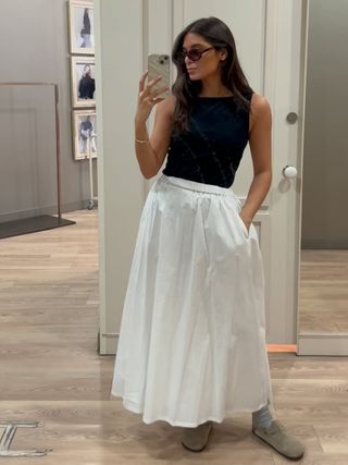 Woman in mirror takes photo wearing black top and white skirt