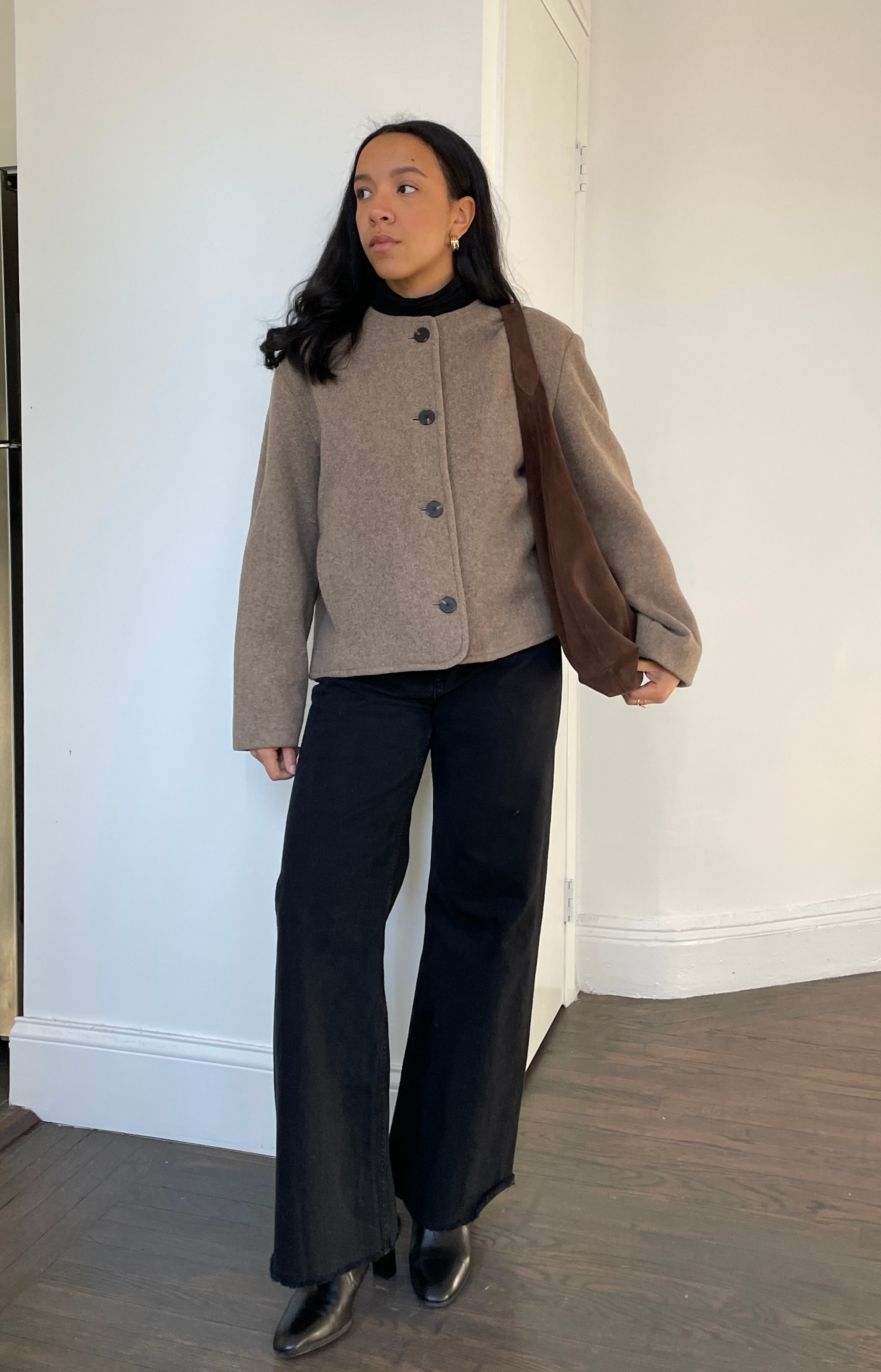 Lauren Tappan wearing a collarless coat, black jeans, black boots, and a brown suede tote