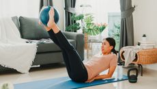 A woman practices Pilates at home on a mat. She is reclining, with her elbows behind her, back touching the mat and feet elevated in the air holding an inflated Pilates ball. Behind her we see a couch and next to her is a small kettlebell.