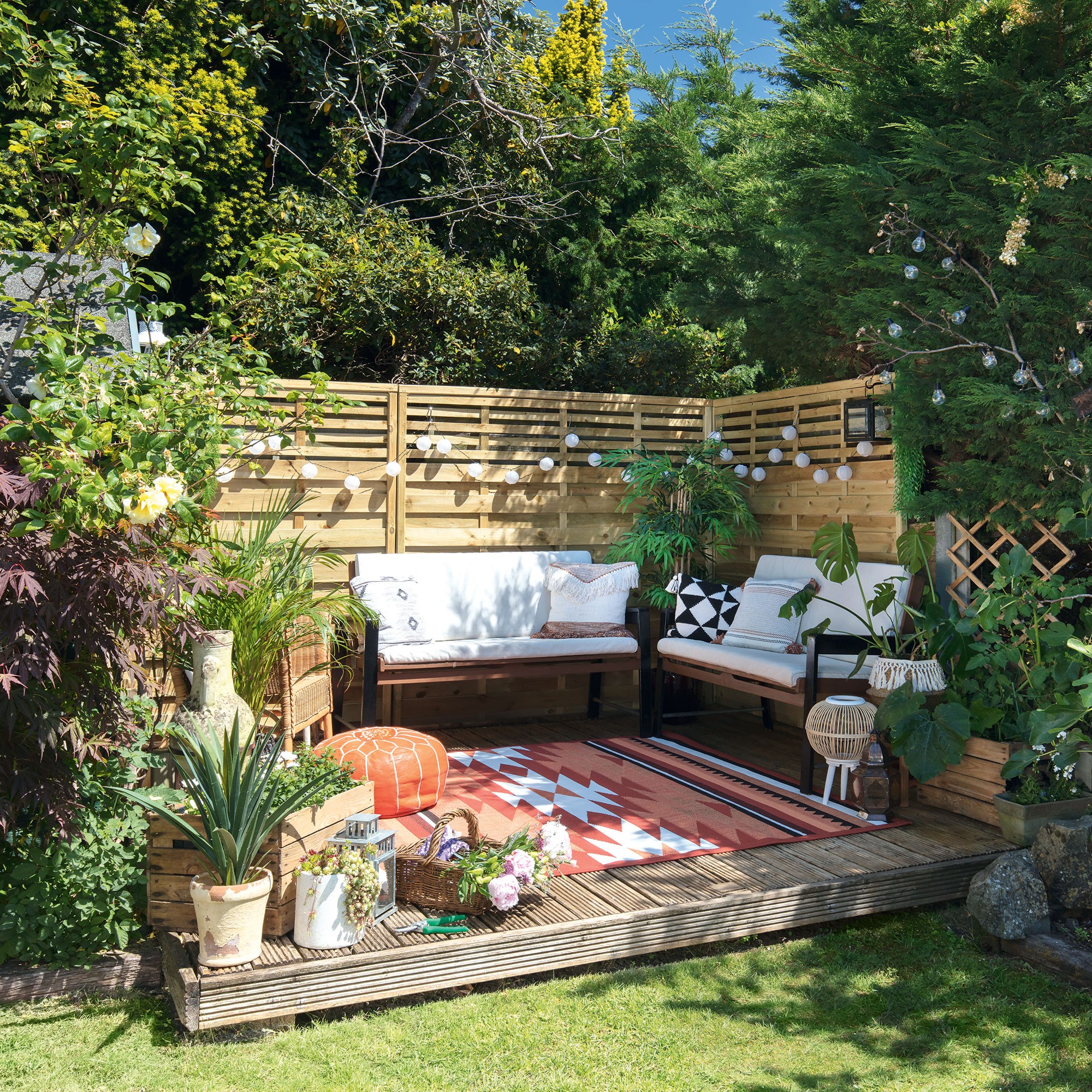 raised decking area in corner of garden with two sofas, patterned rug and potted plants