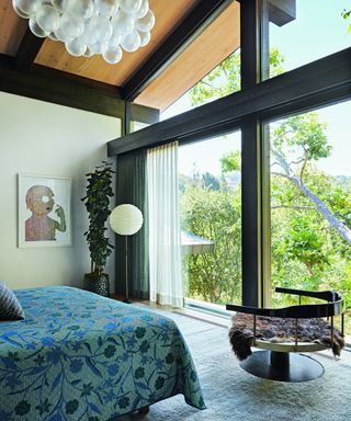 bedroom with cloud-like pendant, bed with blue cover, modern curved chair and floor to ceiling black metal windows