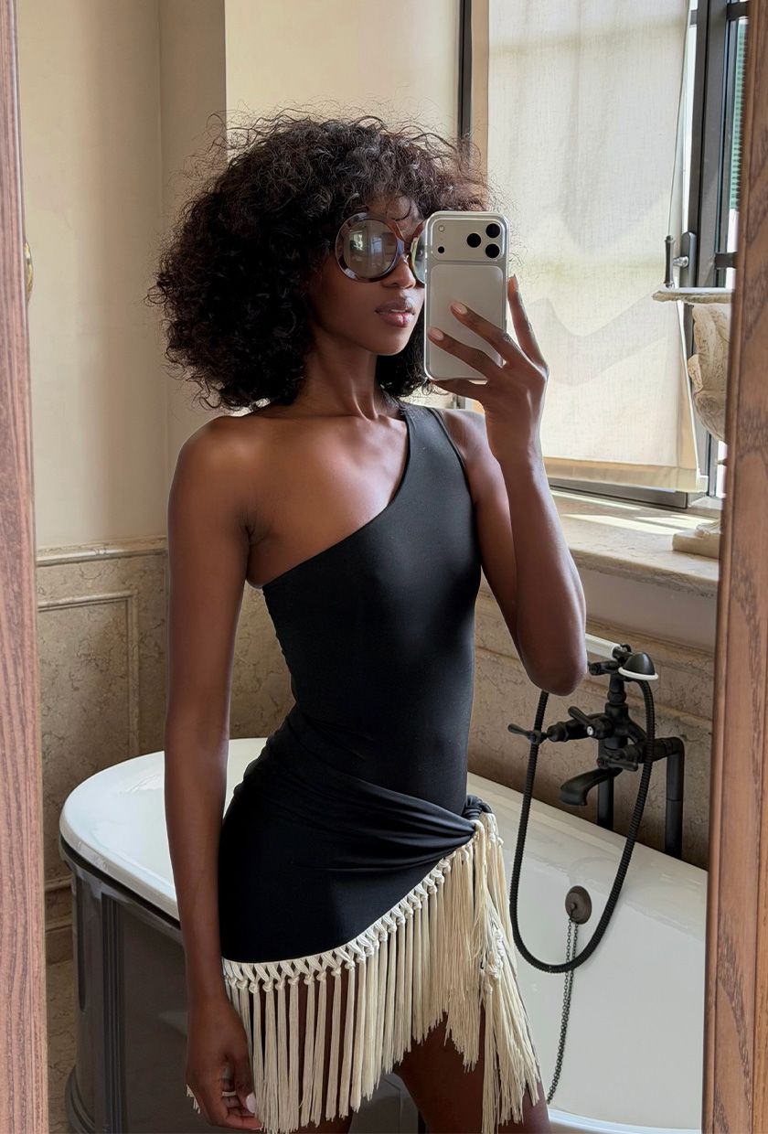 classic swim trend shown on a French woman taking a selfie in the bathroom wearing oversized sunglasses with a black asymmetrical one-piece swimsuit styled with a fringe sarong