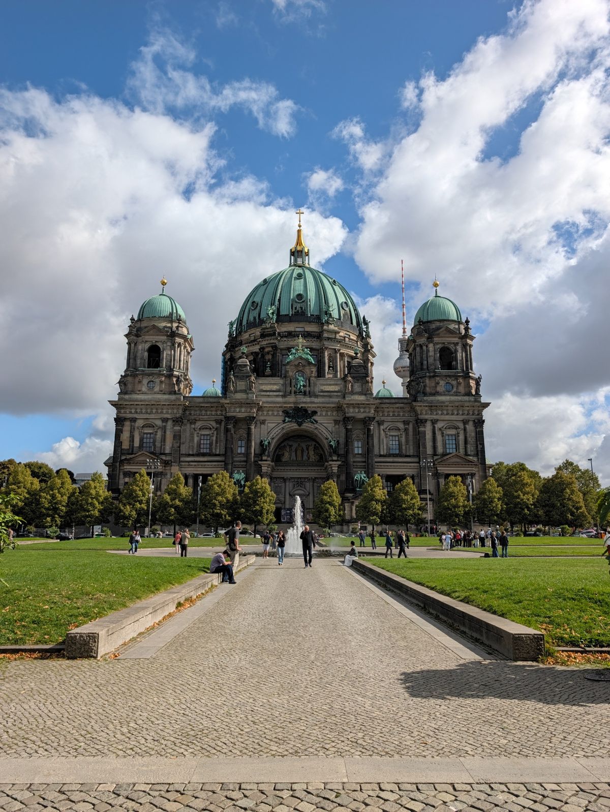 Front view of Berlin Cathedral with its green domes beneath bright clouds, with visitors gathered outside.