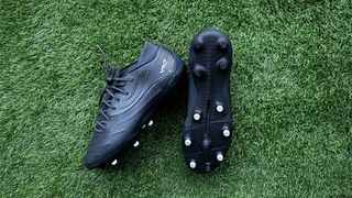 Two black football boots, both with white studs are on green artificial grass. Kipsta Viralto 4 Premium Leather