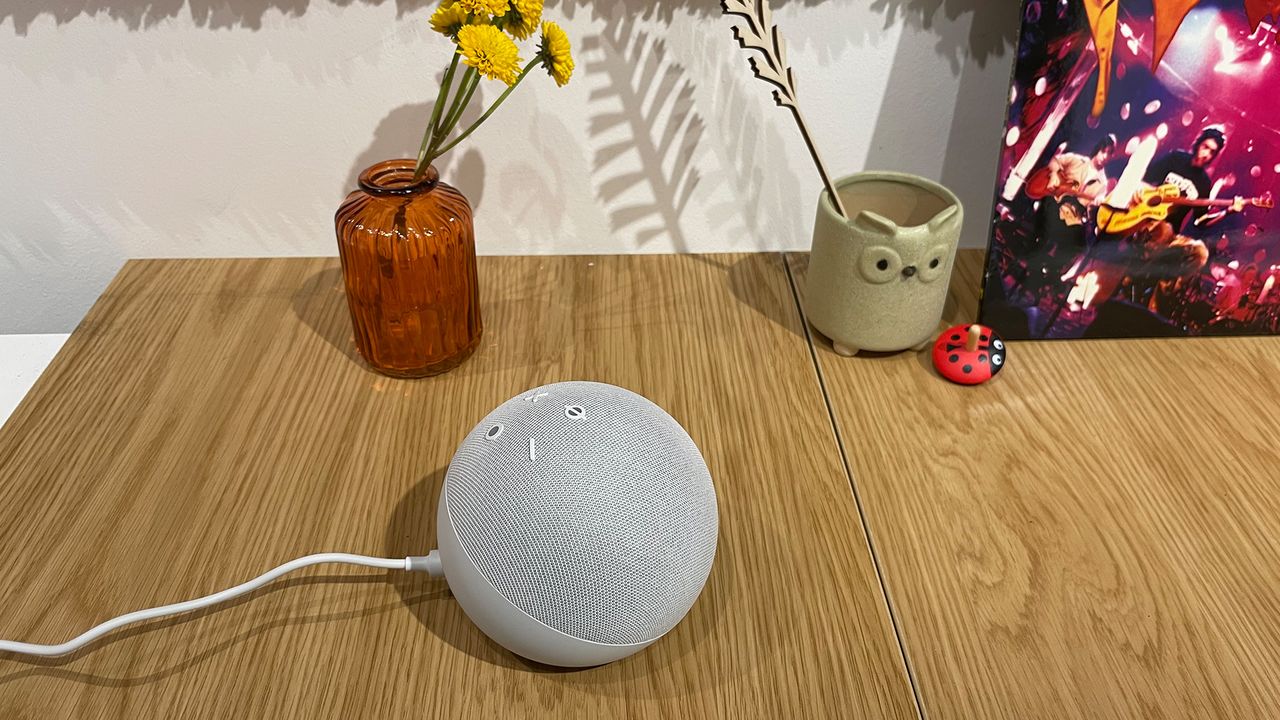 A side view of a white Amazon Echo Dot (5th Generation) smart speaker positioned on a wooden surface. There are some flowers and ornaments in the background.
