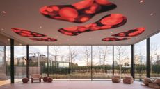 An expansive lounge room with beige-tinted armchairs and sculptural wooden furniture, floor-to-ceiling, wrap-around windows, and an art installation-like ceiling with red organically shaped portholes.