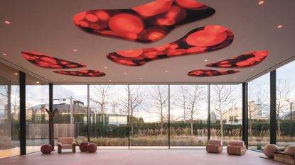 An expansive lounge room with beige-tinted armchairs and sculptural wooden furniture, floor-to-ceiling, wrap-around windows, and an art installation-like ceiling with red organically shaped portholes.