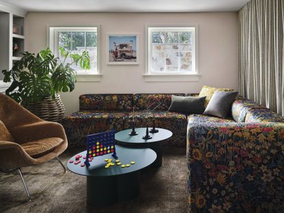 modern living room with pink walls, artwork on wall between two small windows, curtains along one wall, a floral patterned sofa, two nesting coffee tables with Connect Four game, orange velvet armchair, a potted monstera plant and white ceiling with brown rug