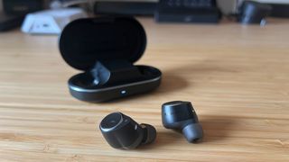 Cambridge Audio M100 earbuds on a wooden desk with charging case in the background