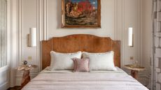 Image of a wooden, classic bedframe, with light gray and dusty pink bedding. The room is a traditional style and there are two marble side tables on either side of the bed with sconce lighting above them.