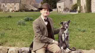 James Anthony-Rose in a brown coat and hat as Carmody sits on a wall with Hamish the dog in All Creatures Great and Small